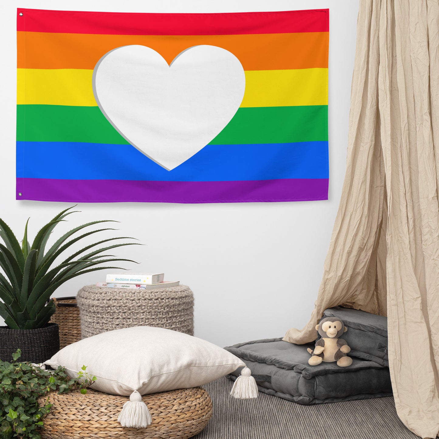 A rainbow pride flag with a large white heart shape in the center, hanging on a wall above a cozy room setup with a pillow, plants, a stool, and a bean bag.