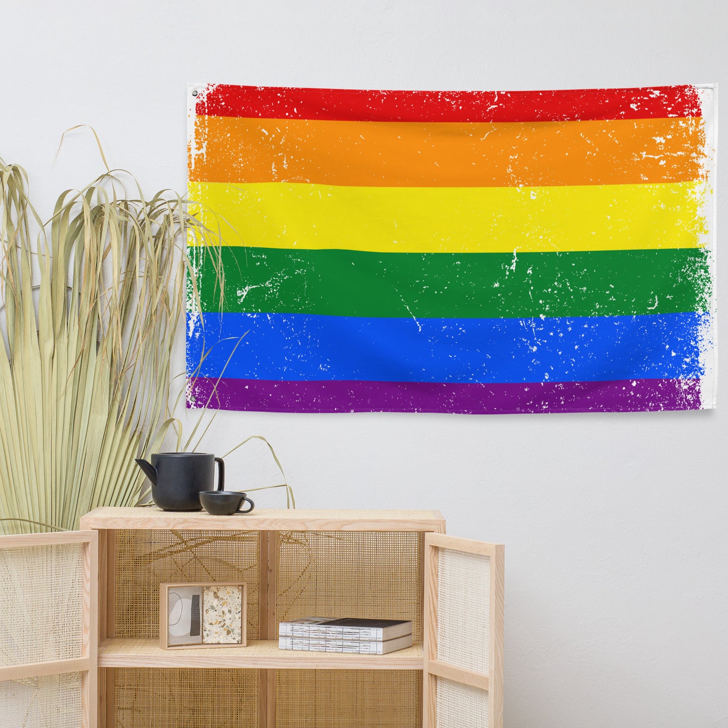 A distressed rainbow pride flag tapestry hangs on a light gray wall above a wooden shelf with decorative items, including a black teapot and cup, and dried grasses on the left.