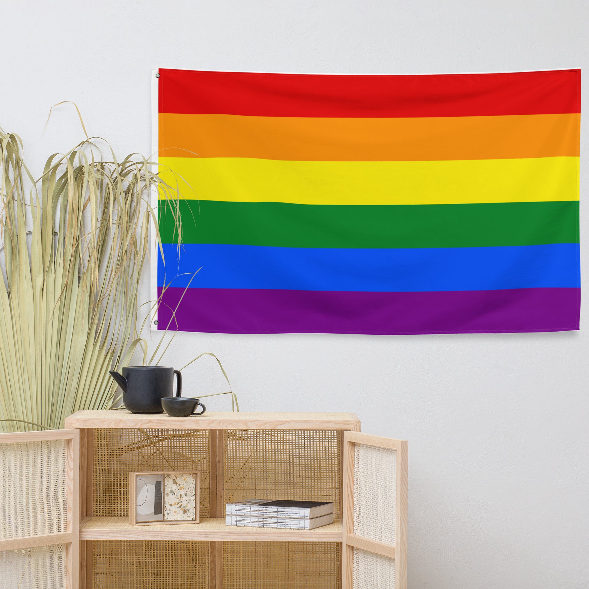 A rainbow pride flag tapestry hanging on a white wall above a light wood cabinet with woven panels, next to dried green plants.