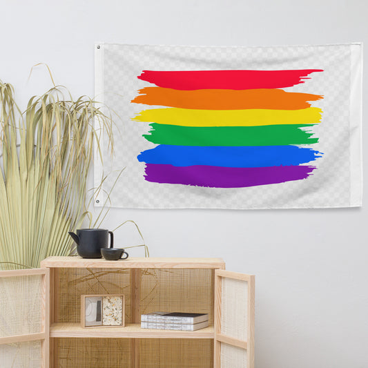 A rectangular wall tapestry with a horizontal rainbow pride stripe pattern hangs on a light gray wall above a wooden cabinet with woven panels. A potted dried plant and a small black teapot and cup sit on the cabinet.