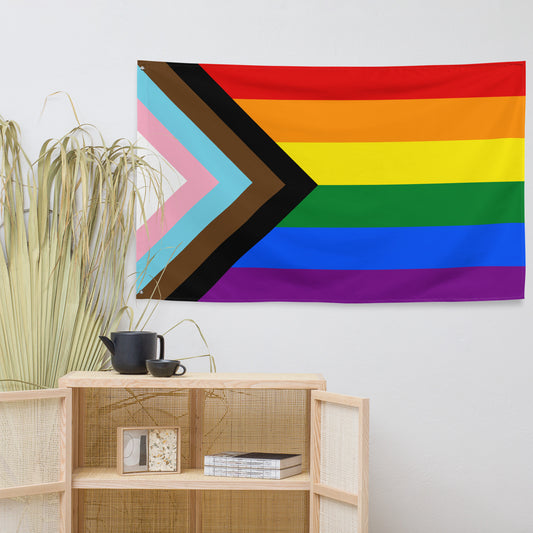 A Progress Pride flag tapestry hanging on a white wall above a light wood cabinet with dried grass stems and a black teapot.