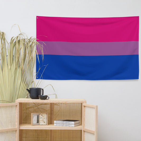 A bisexual pride flag tapestry hanging on a white wall above a light wood shelf with decorative items, including a potted plant.