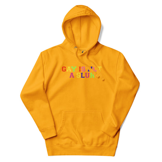 A mustard yellow pullover hoodie with the text 'GAY IS NOT A SLUR' printed across the chest in a rainbow-colored bubble font, displayed on a clean white background.