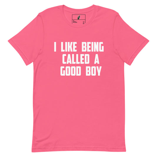 A bright pink short-sleeve t-shirt with bold white text on the front reading 'I LIKE BEING CALLED A GOOD BOY'. The shirt is displayed flat against a solid white background, with a small brand label visible inside the collar.